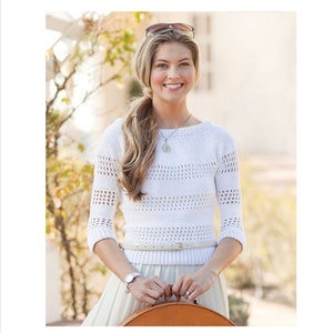 May include: A white, open-weave sweater with three-quarter sleeves. The sweater is paired with a light-coloured skirt and a thin belt. A brown handbag is held in front. Sunglasses are on top of the head.