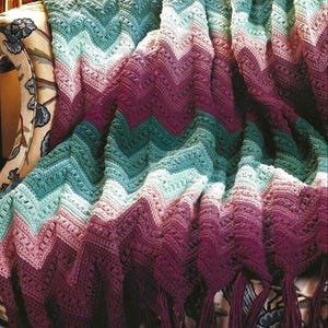 May include: A crocheted throw blanket with a chevron pattern in shades of purple, pink, and green. The blanket has fringe along the bottom edge.