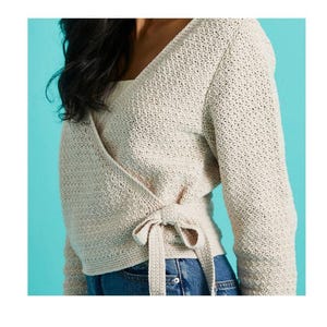 Könnte beinhalten: Cremefarbener Wickelpullover mit langen Ärmeln und Bindeband in der Taille. Der Pullover hat ein strukturiertes, offen gestricktes Design und einen V-Ausschnitt. Die Ärmel sind lang und eng anliegend, kombiniert mit einer blauen Jeans.