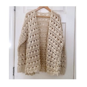 May include: A cream-colored, open-front crochet cardigan with long sleeves. The handmade sweater features a delicate openwork design and ribbed cuffs. The cardigan is hanging on a wooden hanger against a white door.