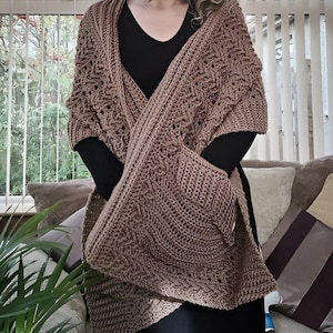 May include: A brown crocheted shawl with a large pocket on the front. The shawl is worn over a black top and black pants.