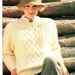 Peut inclure: Un pull en tricot blanc à câble avec un col rond. Le pull est porté par une personne portant un chapeau et un pantalon à carreaux.