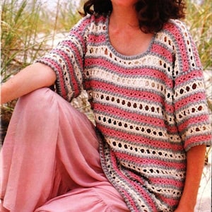 Puede incluir: Una mujer lleva una blusa de crochet a rayas en color rosa y blanco. La blusa tiene un corte holgado y un escote redondo. La mujer está sentada en una playa de arena.