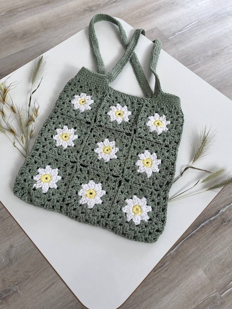 Peut inclure: Un sac cabas en crochet vert avec des fleurs de marguerite blanches et jaunes. Le sac a deux longues anses.