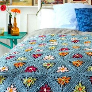 Op de afbeelding: Een blauwe gehaakte deken met een bloemenpatroon. De deken is gemaakt met verschillende kleuren, waaronder roze, oranje, geel en groen. De deken ligt op een bed met witte en blauwe kussens.