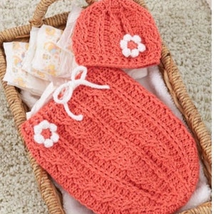 May include: A coral-colored, crocheted baby set, including a hat and a sleep sack, each adorned with a white flower. The set is presented in a woven basket with diapers. The sleep sack has a white bow. The basket is on a light-colored carpet.