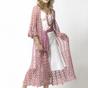 Op de afbeelding: Een lange, roze gehaakte cardigan met een delicaat opengewerkt patroon. De cardigan heeft een gegolfde zoom en driekwartmouwen. Het model draagt een witte jurk en een kralenketting.