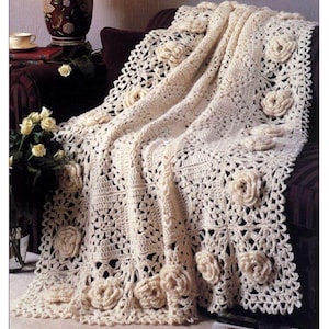 Op de afbeelding: Een gehaakt deken met een delicaat bloemenpatroon. Het deken is gemaakt van wit garen en heeft over de hele oppervlakte verhoogde rozen.