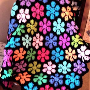 Könnte beinhalten: Eine gehäkelte Decke mit einem sich wiederholenden Blumenmuster. Die Blumen sind in leuchtenden Farben wie Rosa, Blau, Grün und Orange auf schwarzem Hintergrund gehalten. Die Decke ist aus weichem Garn gefertigt.