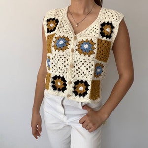 Puede incluir: Un chaleco blanco de ganchillo con un patrón de cuadrados de la abuela. El chaleco tiene cuello en V y es sin mangas. Los cuadrados son en tonos de blanco, marrón, negro y azul.