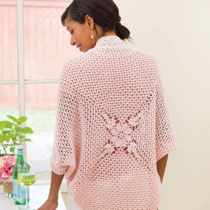 May include: A light pink, open-weave crochet cardigan with short sleeves. The back features a decorative floral design. The cardigan is worn by a person, and the image is taken from behind.