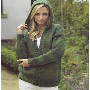 May include: A green hooded knitted sweater with a zip. The sweater is a medium green colour. The model is wearing a white top and white trousers. The background is a garden setting with green foliage and flowers.