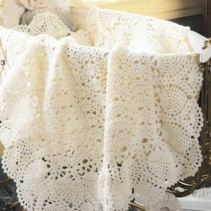 Può includere: Una copertina per neonati in crochet bianco con un delicato motivo a pizzo. La copertina è drappeggiata su una culla e una bambola è sdraiata sopra.