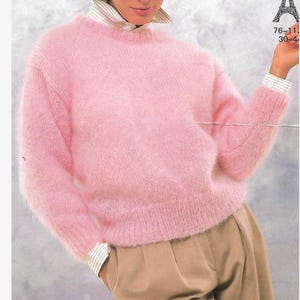 May include: A pink fluffy jumper with a round neckline. The jumper is being worn by a person wearing a white collared shirt and brown trousers.