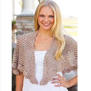 Op de afbeelding: Een beige gehaakte sjaal met een geschulpte rand. Het open weefpatroon is zichtbaar en de sjaal is over de schouders gedrapeerd. Het kledingstuk wordt over een witte top gedragen. De sjaal lijkt handgemaakt.