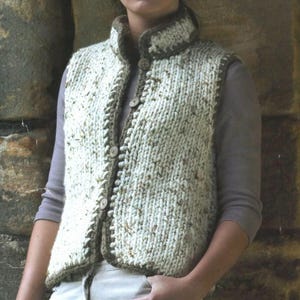 Könnte beinhalten: Eine weiße Strickweste mit braunen Besätzen und Knopfverschlüssen. Die Weste hat einen hohen Kragen und eine gesprenkelte Textur.