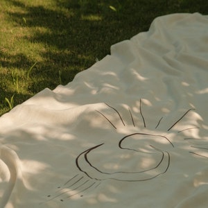 May include: A white linen throw blanket with a black sun design. The blanket is laid out on green grass.