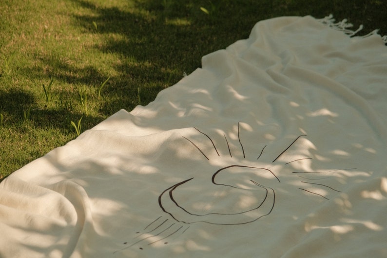 May include: A white linen throw blanket with a black sun design. The blanket is laid out on green grass.