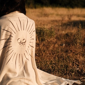 May include: A person sitting in a field wearing a cream-colored blanket with a sunburst design and an Om symbol in the center.
