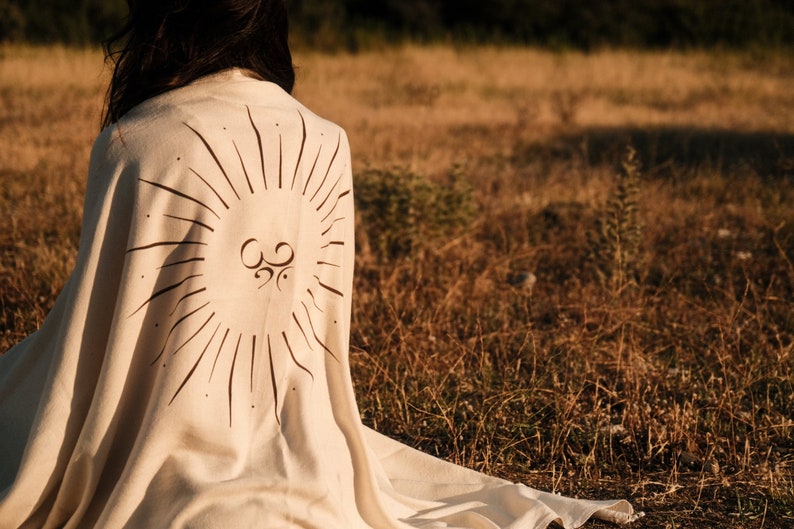 May include: A person sitting in a field wearing a cream-colored blanket with a sunburst design and an Om symbol in the center.