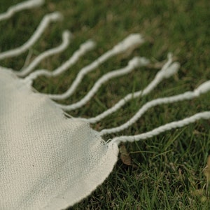 May include: A white fabric with a fringe edge, lying on a bed of green grass.