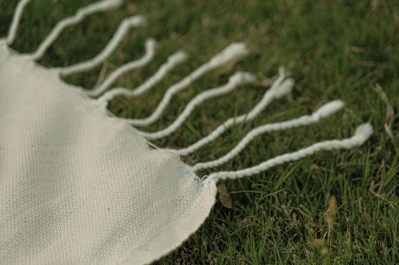May include: A white fabric with a fringe edge, lying on a bed of green grass.