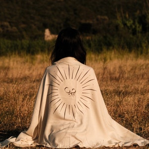 May include: A person sits in a field wearing a white blanket with a sunburst design and the symbol "Om" in the center.