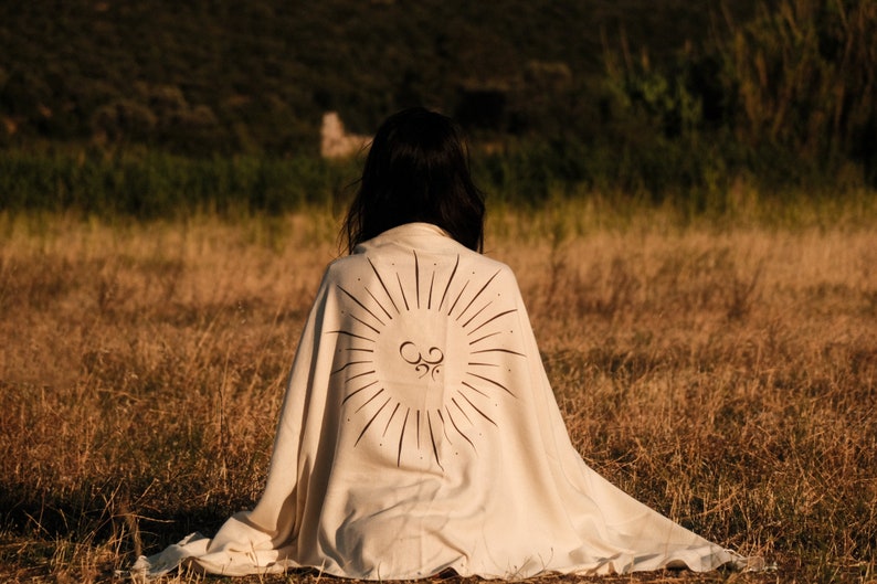 May include: A person sits in a field wearing a white blanket with a sunburst design and the symbol "Om" in the center.