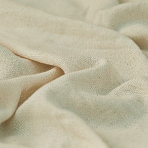 May include: Close-up of a natural-colored fabric with a textured weave. The fabric is folded and draped, showing its soft and drapey texture.
