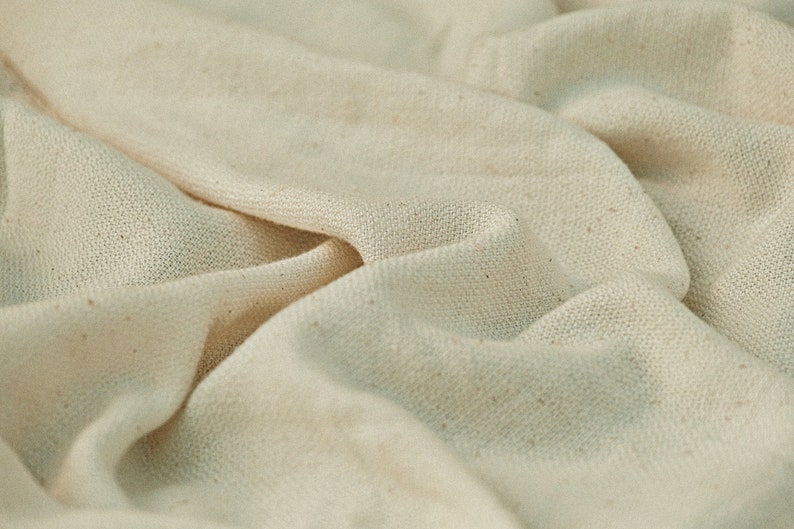 May include: Close-up of a natural-colored fabric with a textured weave. The fabric is folded and draped, showing its soft and drapey texture.