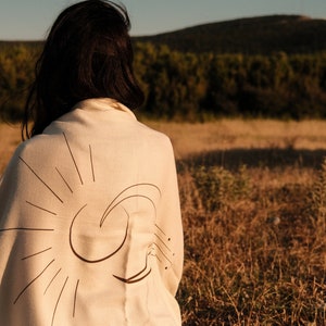 May include: A person wearing a white shawl with a black sun and moon design. The shawl is draped over the person's shoulders and back. The person is standing in a field of tall grass.