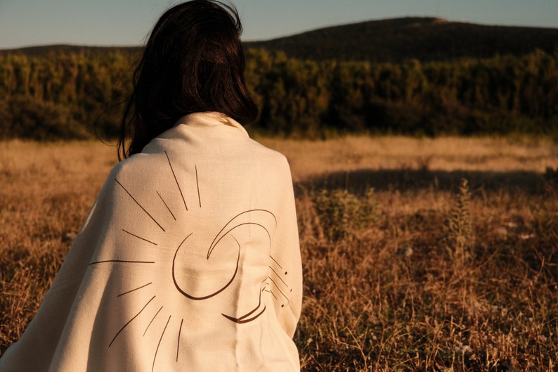 May include: A person wearing a white shawl with a black sun and moon design. The shawl is draped over the person's shoulders and back. The person is standing in a field of tall grass.