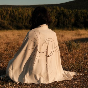 May include: A person sitting in a field wearing a white blanket with a black sun and moon design.