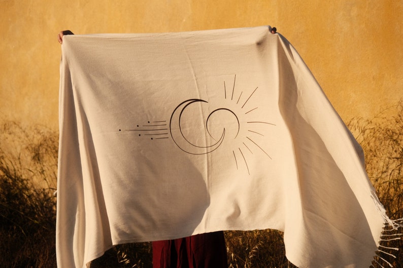 May include: A white cotton throw blanket with a black and white sun and moon design. The blanket has fringe on the edges.