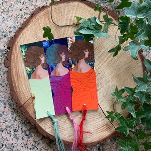 Può includere: Tre segnalibri con un'immagine retrò di una donna su uno sfondo colorato. I segnalibri hanno nappine di colore verde, rosa e rosso.