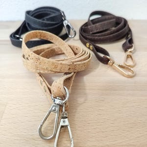Könnte beinhalten: Drei Umhängetaschenriemen in Schwarz, Braun und Kork. Der schwarze Riemen ist aus glattem Material, der braune Riemen hat goldfarbene Beschläge und der Korkriemen hat silberfarbene Beschläge. Alle Riemen haben Metallverschlüsse.