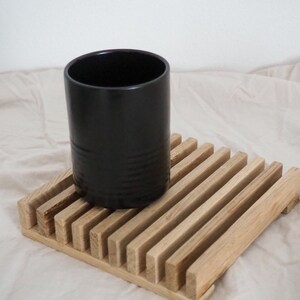 Könnte beinhalten: Eine schwarze zylindrische Keramiktasse steht auf einem Holzuntersetzer mit Lamellen. Die Tasse hat eine glatte, matte Oberfläche und steht auf einem quadratischen Untersetzer aus hellem Holz. Der Untersetzer hat parallele Holzlamellen.