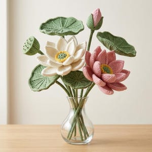 Puede incluir: Un florero de vidrio transparente con un ramo de flores y hojas de loto tejidas a ganchillo. El arreglo incluye un loto blanco, un loto rosa, un capullo de loto rosa cerrado, una vaina de semillas verde y hojas de loto verdes texturizadas.