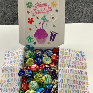 May include: A white gift box with a colourful Happy Birthday card on the lid. The box is open and filled with assorted chocolates wrapped in colourful foil. The box is sitting on a white and pink patterned paper.