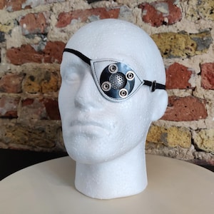 Op de afbeelding: Een zwart-wit ooglapje met een cirkelvormig ontwerp en zilveren klinknagels, gedragen op een witte mannequinhoofd. Het ooglapje heeft een zwarte band en staat tegen een bakstenen muur.