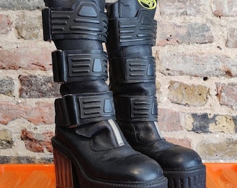 Könnte beinhalten: Schwarze Plateau-Stiefel mit mehreren verstellbaren Riemen und einer klobigen Sohle. Die Stiefel haben einen hohen Schaft und einen metallischen Akzent an der Ferse. Der Markenname "Tracker's" ist in gelber Schrift auf der Oberseite der Stiefel zu sehen.
