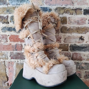 Puede incluir: Un par de botas de plataforma beige con ribete de piel sintética. Las botas tienen un diseño con cordones y una suela gruesa. Las botas están sobre un fondo de pared de ladrillo.