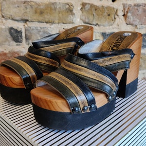 Può includere: Coppia di zoccoli platform in pelle nera e marrone con tacchi in legno. Gli zoccoli hanno un design a righe e sono decorati con borchie metalliche.