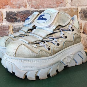 Puede incluir: Un par de zapatillas de plataforma blanquecinas con múltiples correas y una suela gruesa. Las zapatillas tienen un logotipo en la lengüeta y están hechas de un material texturizado. Los zapatos están sobre una superficie verde.