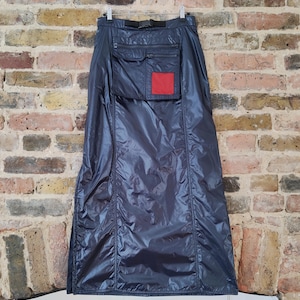May include: A long, navy blue skirt made of a shiny, water-resistant material. The skirt features a large pocket with a red square patch and a black belt. The skirt is hanging on a hanger against a brick wall.