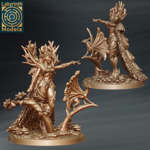 May include: Two golden figurines of a female figure with wings and a small bird. The figures are standing on a base with a leafy design. The figures are both wearing elaborate clothing and jewelry.