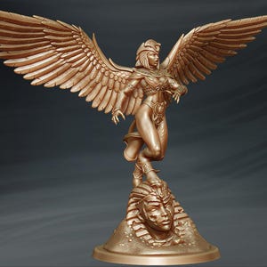 May include: A gold-coloured statue of a winged woman with a headdress standing on a base with a sculpted head. The woman is holding her arms out in front of her.