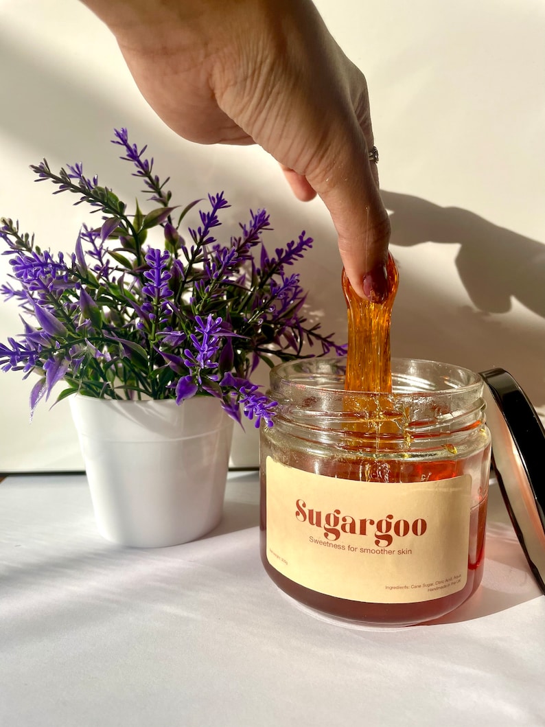 May include: A glass jar of amber-colored sugar wax with a black lid. The jar has a label that reads "Sugargoo Sweetness for smoother skin".