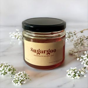 May include: A glass jar with a brown label that reads "Sugargoo Sweetness for smoother skin". The jar is filled with a light brown liquid and has a black lid.
