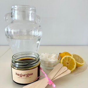 May include: A brown jar of sugar wax labeled "SugarGoo" with the tagline "Sweetness for smoother skin".  The jar is next to a bowl of white sugar cubes, a plate with three lemon wedges, and three wooden sticks tied together with a pink ribbon.